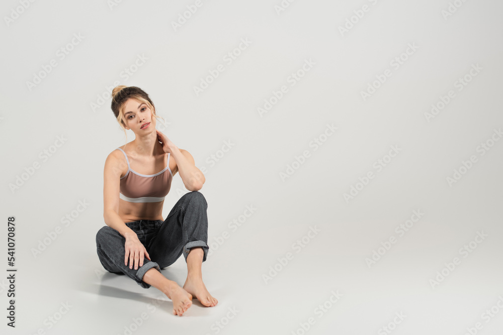 pretty barefoot woman with slim body sitting in jeans and sports top on grey background pretty barefoot woman with slim body sitting in jeans and sports top on grey background