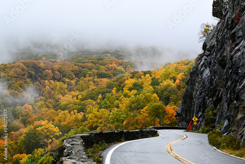 A mountain road in Fall carves into the side of a mountain in front of the edge of the Hudson River and a large forest disappearing into the fog.