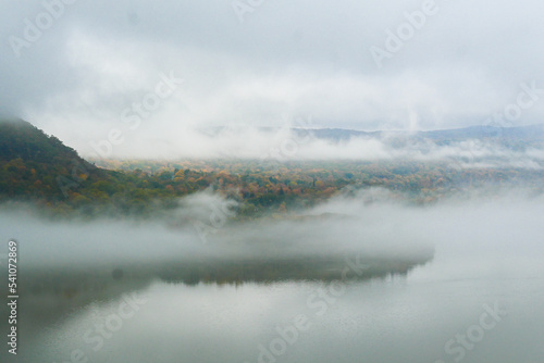 Forests and mountains of the Hudson River Valley break through the fog and reflect over the water during fall foliage.