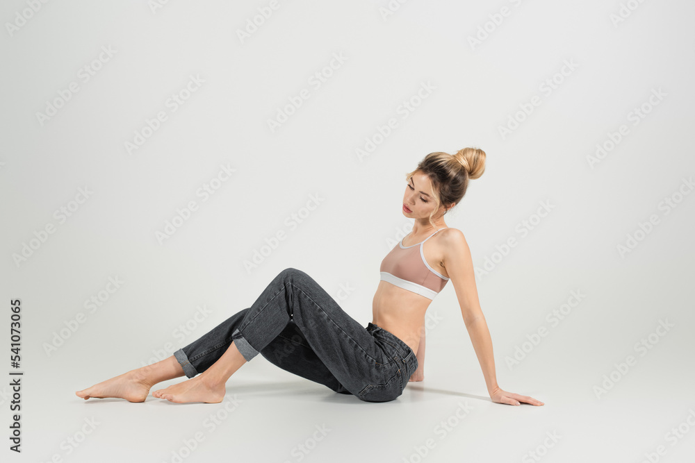 full length of young barefoot woman with perfect figure sitting in jeans on grey background full length of young barefoot woman with perfect figure sitting in jeans on grey background