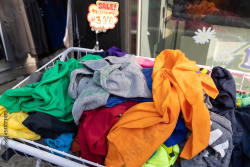 A shopping basket filled with clothes with a note: Sale, cash only. Bunch of t-shirts of different colors for sale. Cheep clothing and fast fashion theme