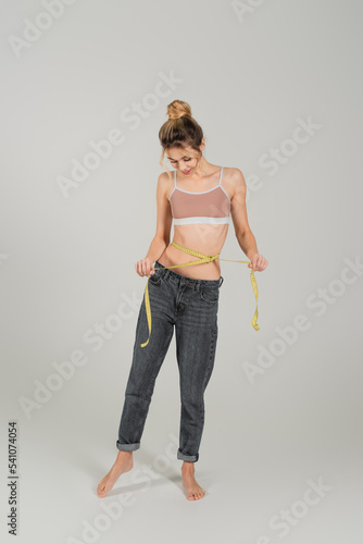 full length of barefoot woman in jeans and sports top measuring waist on grey background
