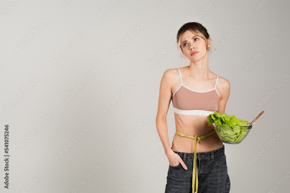 upset woman with measuring tape on waist holding bowl of fresh lettuce isolated on grey upset woman with measuring tape on waist holding bowl of fresh lettuce isolated on grey