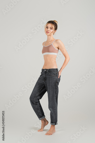 full length of fit woman in sports top and jeans standing barefoot on grey background © LIGHTFIELD STUDIOS