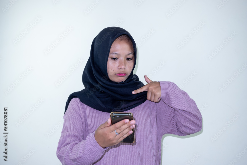 Young Asian woman wearing hijab holding phone in hand and pointing at it with playful expression isolated on white background Young Asian woman wearing hijab holding phone in hand and pointing at it with playful expression isolated on white background