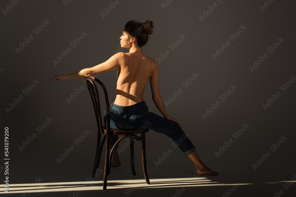 back view of half nude barefoot woman in jeans posing on wooden chair on grey background with lighting back view of half nude barefoot woman in jeans posing on wooden chair on grey background with lighting