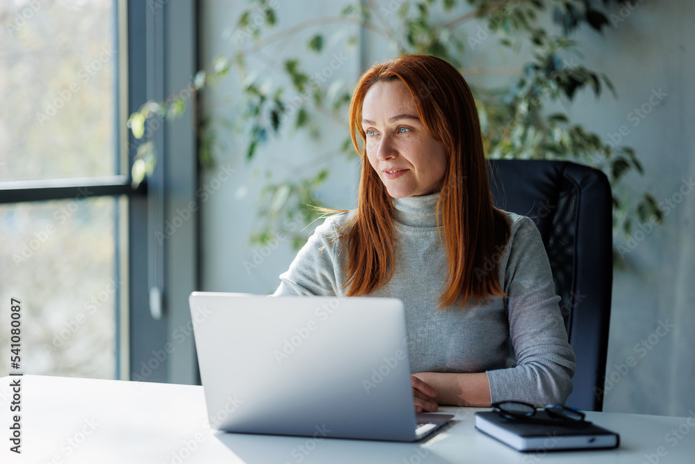 a mature, successful businesswoman seated at a desk in an office in casual attire with her arms crossed. narrow focus a mature, successful businesswoman seated at a desk in an office in casual attire with her arms crossed. narrow focus