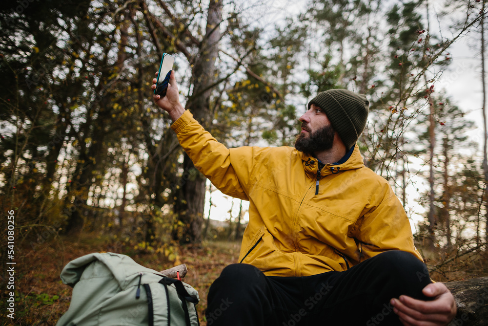 A bearded man rests on a fallen tree in the middle of the forest. A man uses a mobile phone to search GPS.