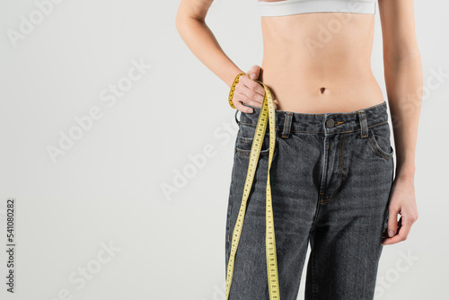 cropped view of fit woman in jeans standing with hand on hip and measuring tape isolated on grey