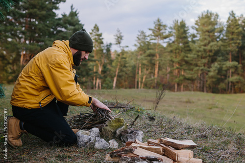 Survival in the wild. A bearded man lights a fire near a makeshift shelter made of pine branches.