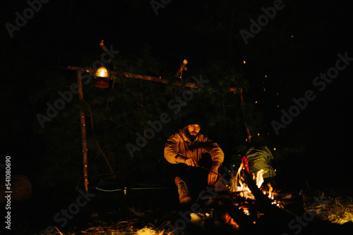 Fototapeta Naklejka Na Ścianę i Meble -  The traveler rests by the night campfire. Furnished shelter from branches for sleeping with a kerosene lamp and a fire surrounded by stones.