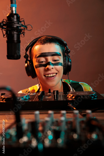 Crazy female artist acting silly at mixer, looking closely at control buttons to mix and remix music wearing headphones. Producing techno sounds with turntables, funky focused disc jockey. photo