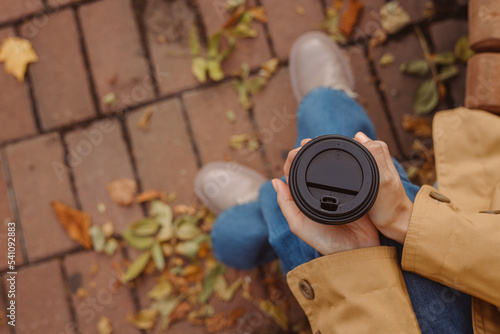 Top view of crop woman holding paper cup of takeaway coffee and warming hands on bench in autumn in park 