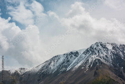 Awesome landscape with high snowy mountain range with sharp rocks in cloudy sky. Dramatic view to snow mountains in changeable weather. Atmospheric mountain scenery with white snow on black rocks.