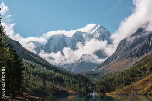 Scenic landscape with turquoise mountain lake in autumn valley against large snow mountains in low clouds in morning sunlight. Alpine lake with view to sunlit high snowy mountain range in low clouds.