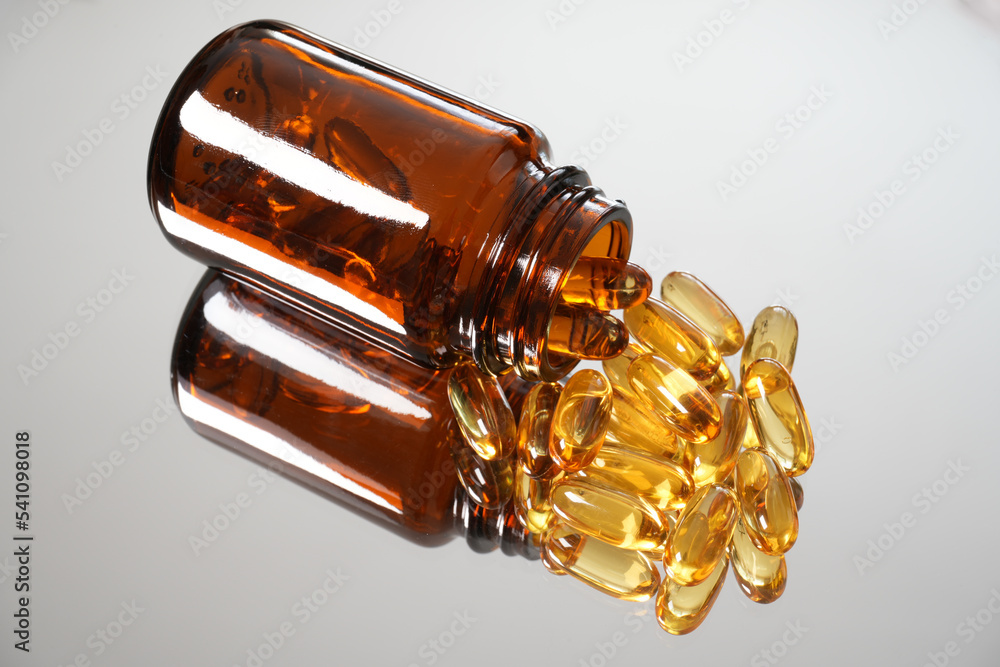 Fototapeta premium Fish oil capsules close-up with selective focus and reflection on a smooth mirror surface. Scattered from brown glass bottle and the half bottle include in screen. Capsules and bottle top-right view.