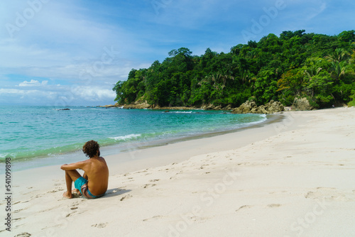 Unrecognizable young man sitting on white sanded beach with jungle in the background.