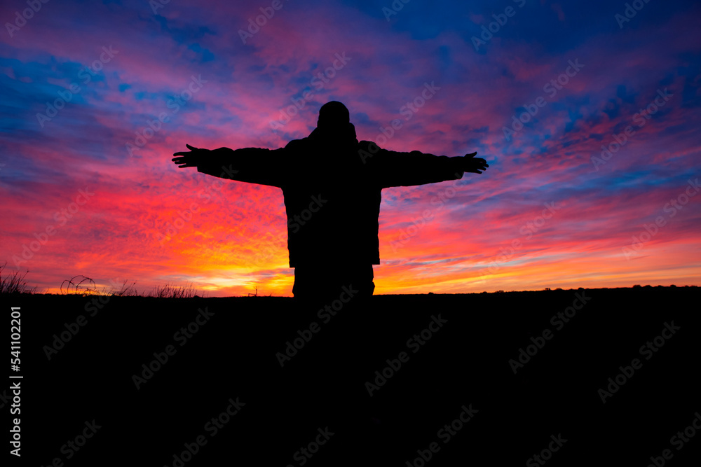 A man with outstretched arms silhouetted against a vibrant desert sunrise.