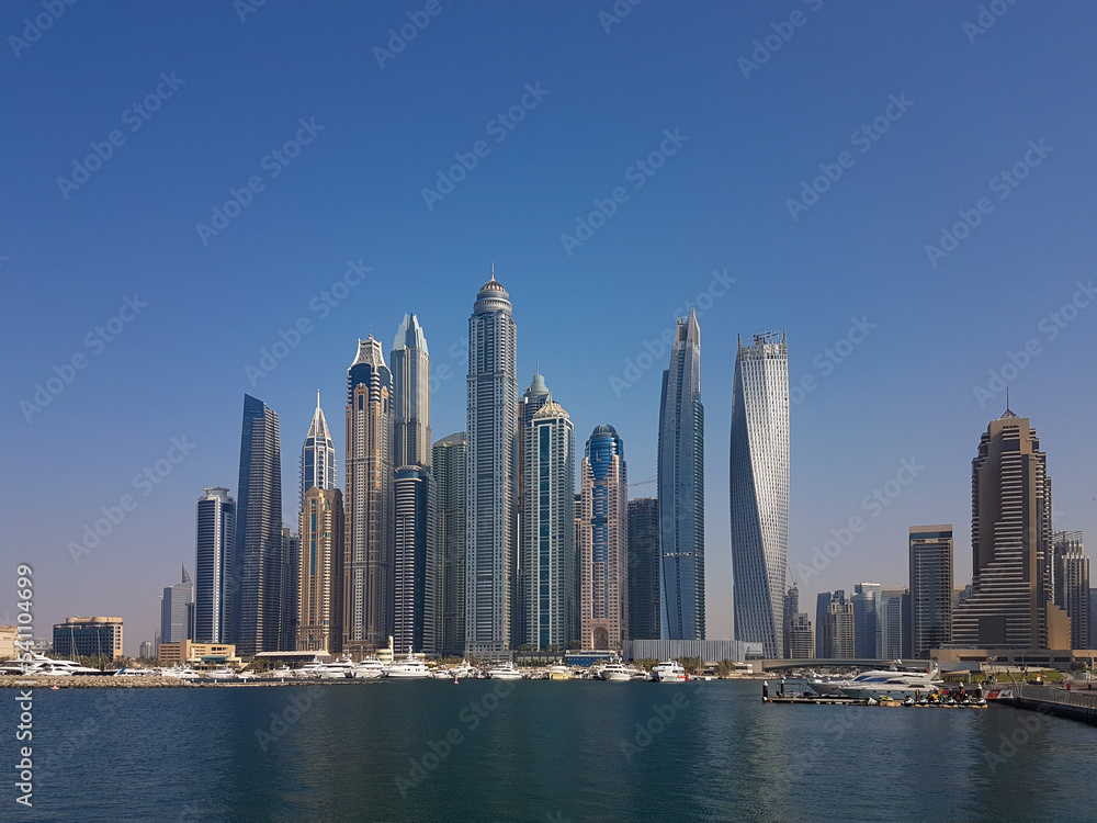 Fototapeta premium Cityscape of tall, modern buildings. Skyscrapers in Dubai Marina on a bright summer day. Clear, blue skies, calm waters, and yachts moored on the docks.