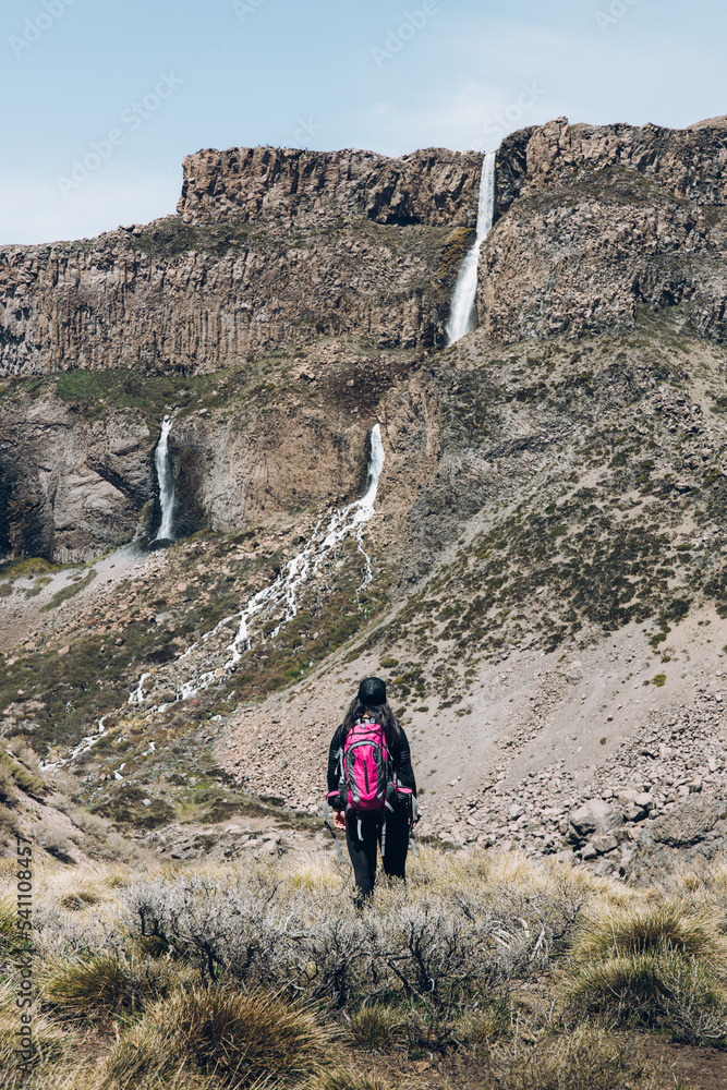 Rear view of female backpacker hiking through a rocky path to a big waterfall. Vertical shot