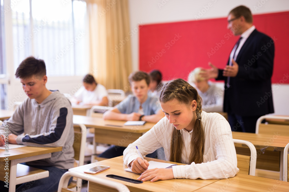 Diligent high school student teenage girl studying in college with classmates, making notes of teacher lecture