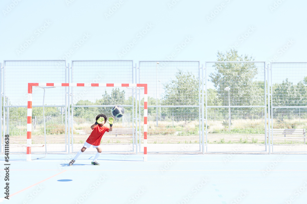 Young afro American boy goalkeeper with red football shirt playing football in a training field. Saving a penalty kick.