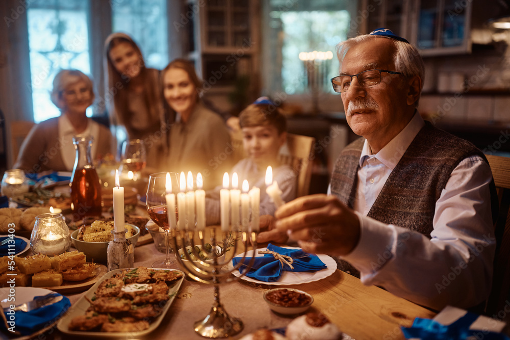Close up of senior Jewish man lights menorah during family dinner on Hanukkah.