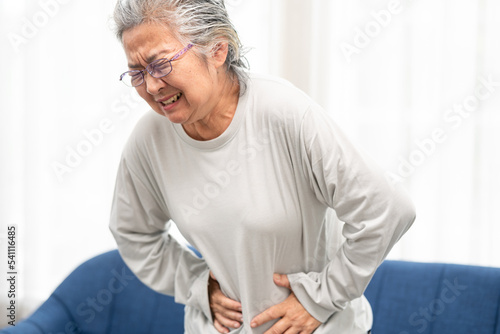 Senior woman suffering from stomach ache at home. Grabbing and squeezing belly with hand, feeling exhausted, standing in living room. Expressing pain on face