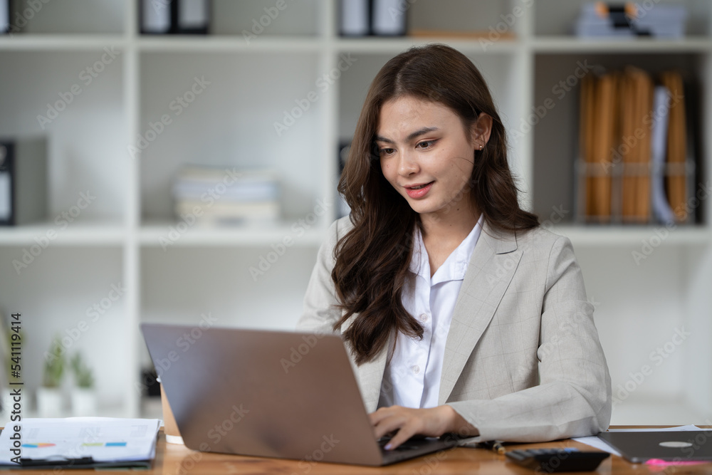 Happy businesswoman typing on laptop at her desk looking at camera in bright office Happy businesswoman typing on laptop at her desk looking at camera in bright office