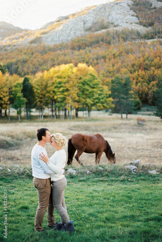 Man kisses woman on the forehead on a green lawn next to a grazing horse