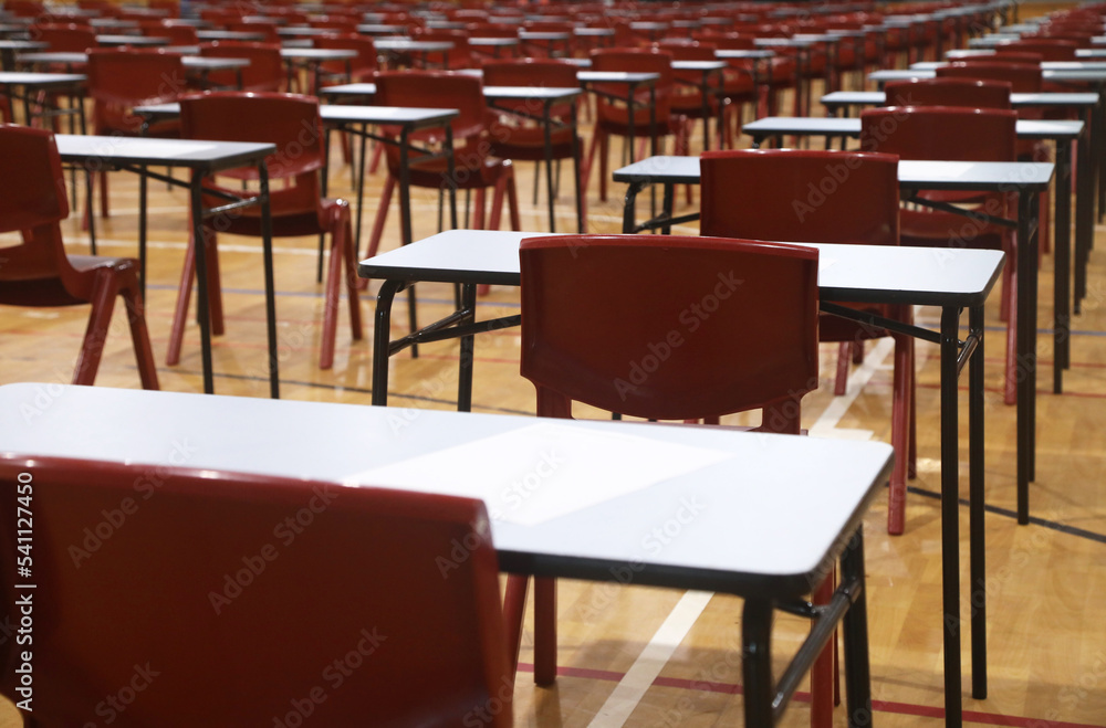 A large school hall filled with multiple exam tables and red chairs set up ready for major exams or student testing.  Rows of examination tables and chairs. Educational concept