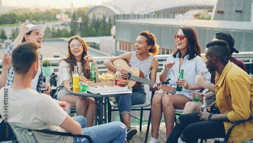Pretty African American girl is playing the guitar while her happy friends multiethnic group are singing and moving hands enjoying party on rooftop. Table with food and drinks is visible.