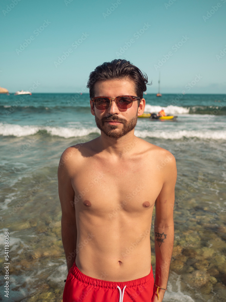 Shirtless young male smiling into the camera in front of the blue beach sea.