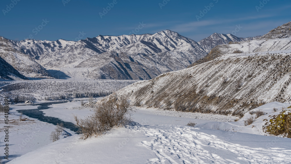 Winter river in a snowy valley. Steam is visible above the unfrozen part of the riverbed. Footprints in snowdrifts. Picturesque mountains against the sky. Altai. Katun. Winter river in a snowy valley. Steam is visible above the unfrozen part of the riverbed. Footprints in snowdrifts. Picturesque mountains against the sky. Altai. Katun.