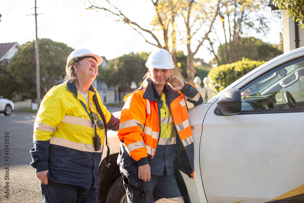 Two smiling women road workers with white helmet leaning on a white car Two smiling women road workers with white helmet leaning on a white car