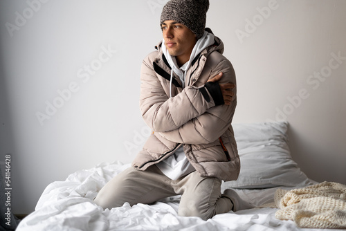 The man in the bedroom wearing wool hat, no heating and electric at home. Men warming in the blanket. Heating season. Cold frozen young man in the room