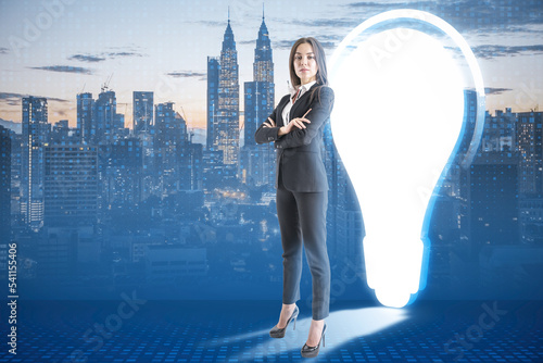Attractive young european businesswoman with folded amrs standing on blurry night city background with abstract glowing light bulb door. Idea, innovation, solution and success concept. © Who is Danny