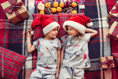 Top view of kids in christmas pajamas and Santa's hats lying on checkered blanket and pillows 