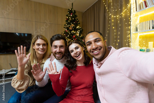 Diverse friends celebrating christmas and new year together  group of people having dinner at home sitting on sofa near Christmas tree  taking selfie photo and talking with friends on video call using