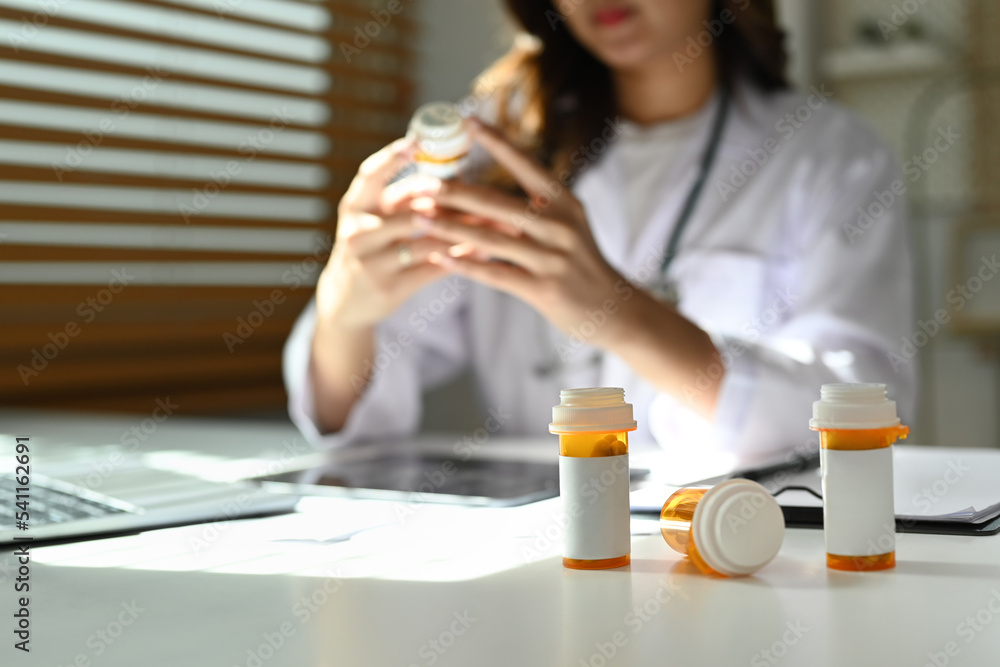 Close up view of medical pill bottles with blank label on white table and female doctor sitting in background Close up view of medical pill bottles with blank label on white table and female doctor sitting in background