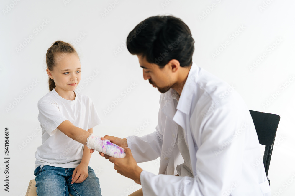 Studio shot of male doctor in uniform consulting little girl patient with broken hand wrapped in plaster bandage at checkup meeting on white isolated background. Concept of insurance and healthcare. Studio shot of male doctor in uniform consulting little girl patient with broken hand wrapped in plaster bandage at checkup meeting on white isolated background. Concept of insurance and healthcare.