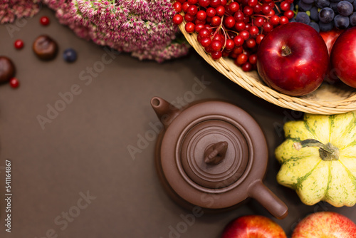 Autumn composition with a harvest of pumpkins, apples, red berries, grapes, flowers. Brown clay teapot on a brown background with copy space. Tea party, autumn, autumn harvest, flowers. flat lay