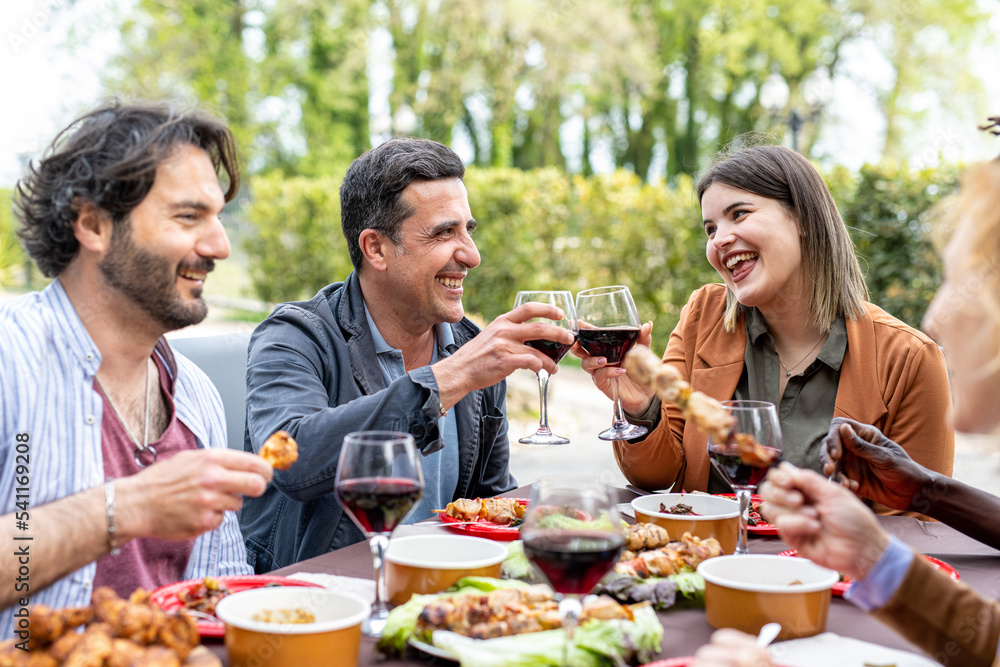 Happy family cheering with red wine at barbecue lunch outdoor, different age of people having fun at weekend meal, food, taste and family concept, focus on smiling young woman