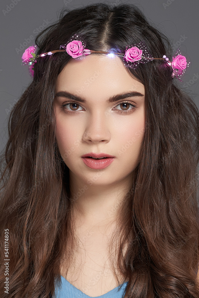 Naklejka premium Cropped close-up shot of a brown-haired young woman with a LED luminous flower wreath on her head. A girl with a pink roses wreath headband is on a gray background. Front view.