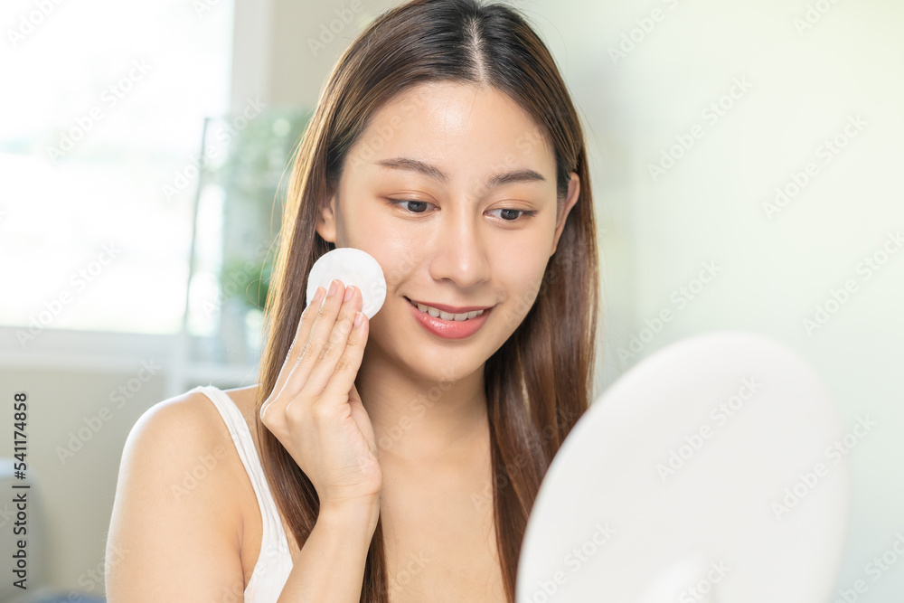 Happy beauty, beautiful asian young woman, girl smiling, looking in mirror, holding cotton pad, applying lotion by wipe on her face, removing makeup before shower at home. Skin care routine people. Happy beauty, beautiful asian young woman, girl smiling, looking in mirror, holding cotton pad, applying lotion by wipe on her face, removing makeup before shower at home. Skin care routine people.