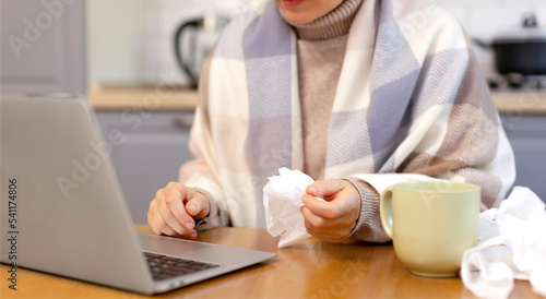 A woman sits in front of a laptop in the kitchen, wrapped in a scarf, holding a napkin in her hand. The woman is sick and working remotely from the office. Napkins, runny nose, flu, orvi, sick leave photo