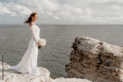 Romantic bride, blond girl in white wedding dress with open shoulders posing with sea and rocks in background. Stylish young woman standing on cliff's edge and looking into a ocean mountains at sunset