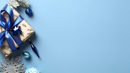 Christmas gift box with blue ribbon bow, snowy fir branches, ornaments on pastel blue background. Flat lay, top view.