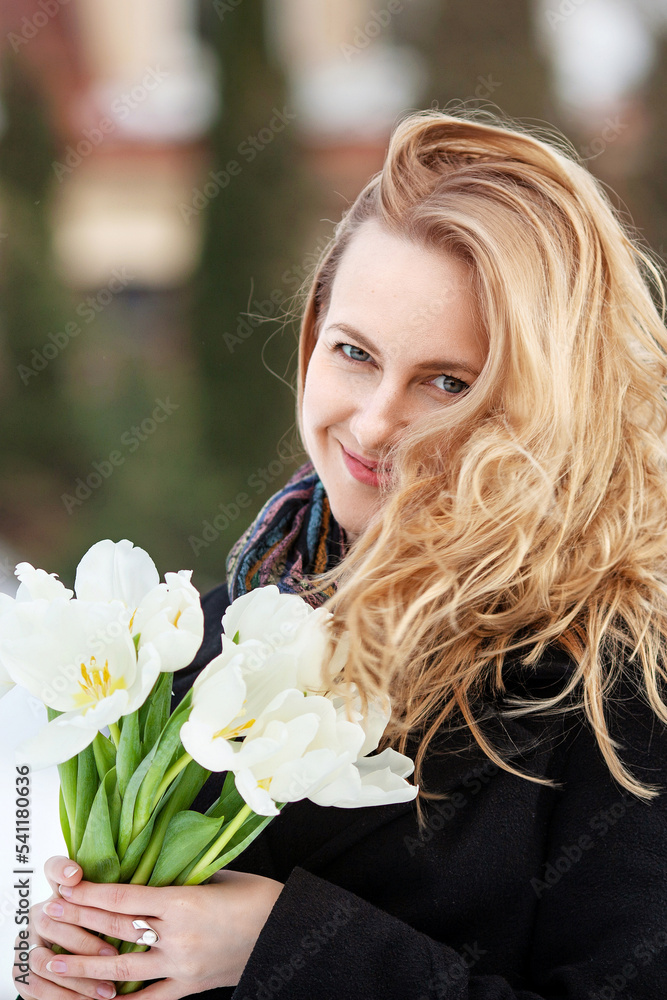 Naklejka premium Elegant smiling young woman with long blonde hair in a black coat poses in the city. Portrait of a beautiful modern woman with flowers