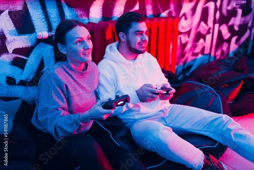 Young man and woman couple plays on game console with gamepad sitting on chair.